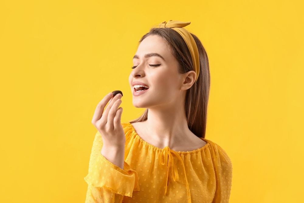 Auf dem Bild sieht man eine Frau vor gelbem Hintergrund, die kurz davor ist, in eine Praline zu beißen. Sie trägt gelbe Kleidung.
