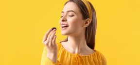 Auf dem Bild sieht man eine Frau vor gelbem Hintergrund, die kurz davor ist, in eine Praline zu beißen. Sie trägt gelbe Kleidung.