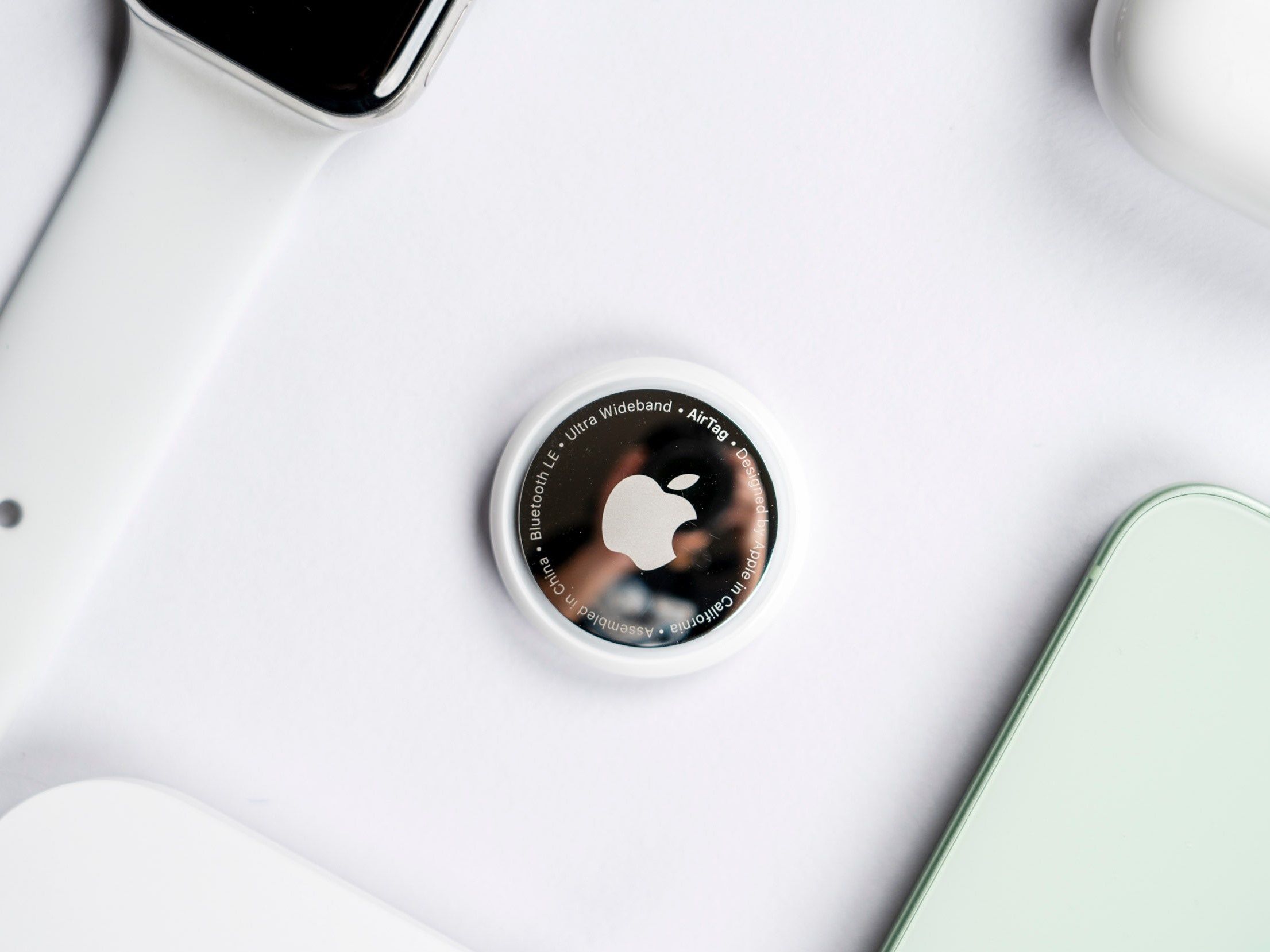 Auf dem Bild sind Apple-Produkte zu sehen (manche nur zum Teil): eine Apple Watch, ein iPhone, AirPods und in der Mitte liegt ein Apple AirTag (Ortungsgerät).
