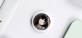 Auf dem Bild sind Apple-Produkte zu sehen (manche nur zum Teil): eine Apple Watch, ein iPhone, AirPods und in der Mitte liegt ein Apple AirTag (Ortungsgerät).